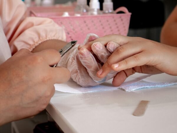 Profissional de manicure usando luvas descartáveis nitrílicas cortando a cutícula de uma cliente com alicate de inox. Foco na biossegurança e nos acessórios de saúde.